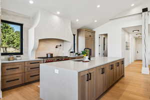 Kitchen with brown cabinetry, light wood-type flooring, recessed lighting, a kitchen island with sink, and light stone countertops