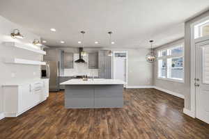 Kitchen with gray cabinets, stainless steel fridge, hanging lights, open shelves, and an island with sink