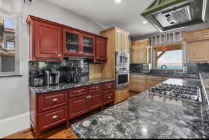 Kitchen featuring exhaust hood, dark stone counters, glass insert cabinets, stainless steel appliances, and recessed lighting