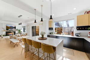 Dual tone kitchen featuring light wood finished floors, a kitchen bar, hanging light fixtures, a glass covered fireplace, and light stone counters
