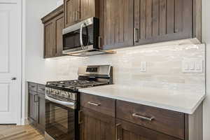 Kitchen featuring stainless steel appliances, tasteful backsplash, dark wood finish cabinets, light stone countertops, and light wood-style floors