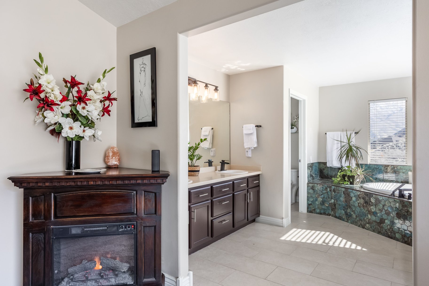 Bathroom featuring a corner electric fireplace, a double vanity with white countertops, and a built-in tub with textured blue-green tile surround