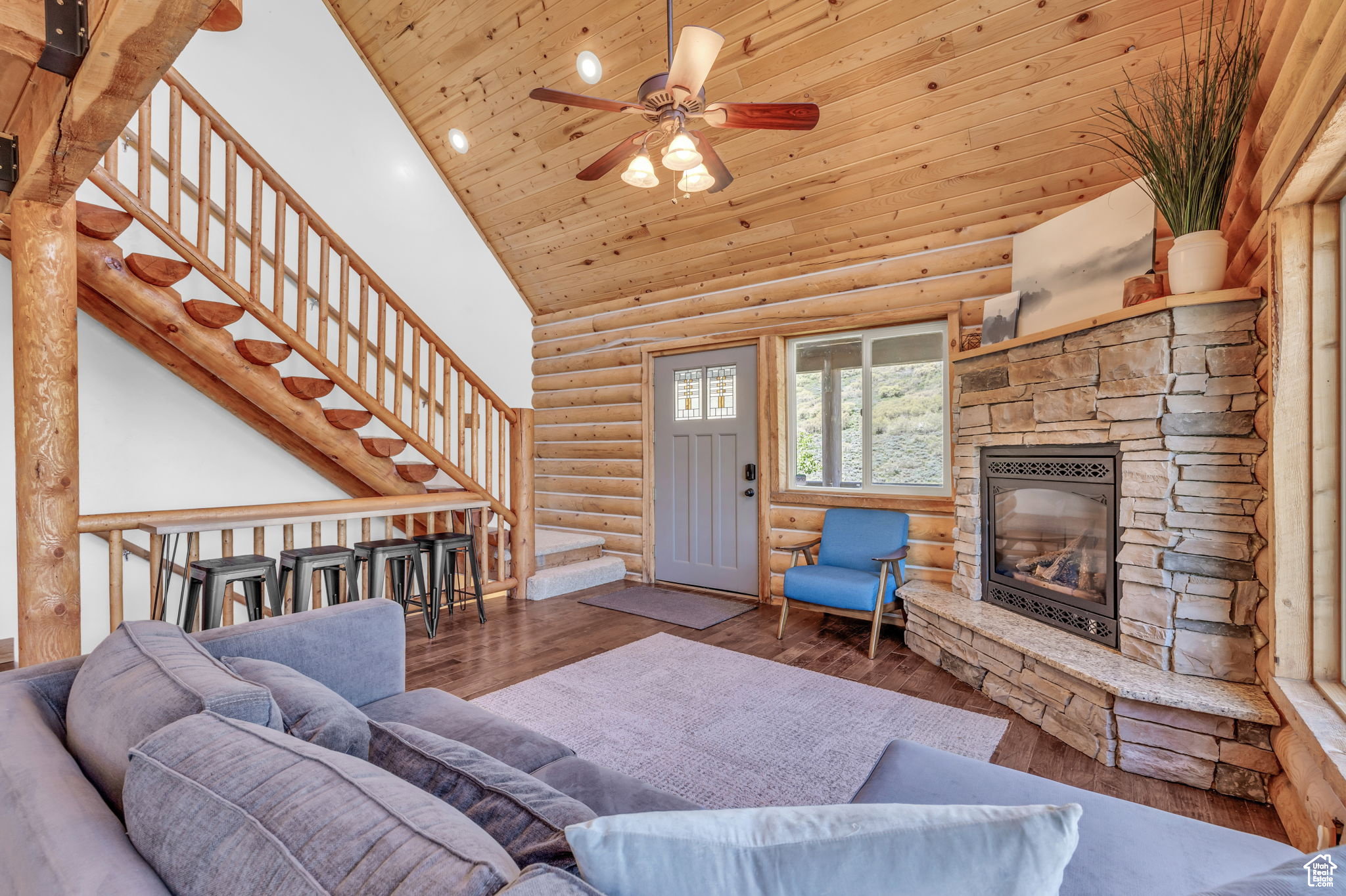 Living room featuring hardwood / wood-style floors, a vaulted wooden ceiling, a stone fireplace, log walls, and ceiling fan