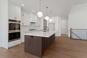 Kitchen with dual tone cabinetry, an island with sink, stainless steel double oven, light wood-type flooring, and decorative light fixtures