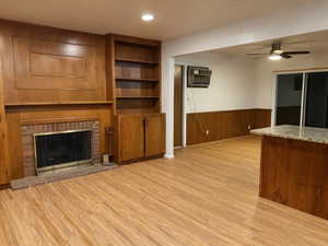 Unfurnished living room featuring wood walls, wainscoting, ceiling fan, light wood-style floors, and a brick fireplace