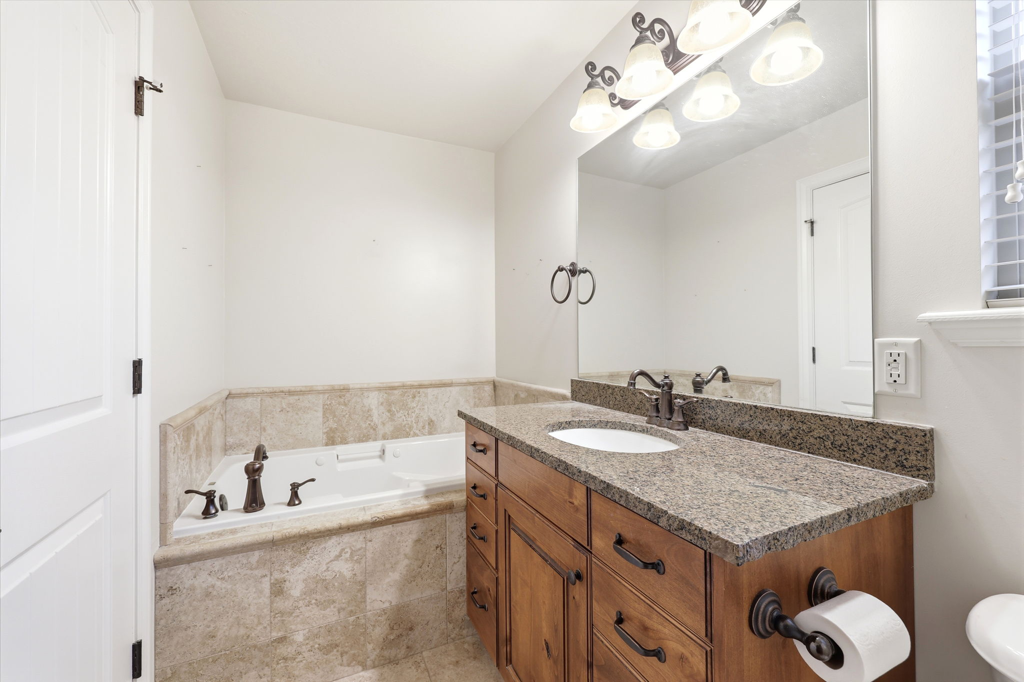 Bathroom featuring an integrated soaking tub with tile surround, a single vanity with wood cabinetry, a stone countertop, an undermount sink, and dark bronze fixtures