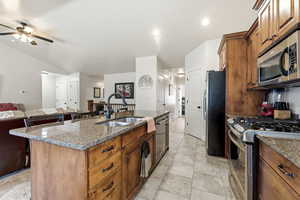 Kitchen featuring wood finish cabinets, stainless steel appliances, light stone counters, a kitchen island with sink, and ceiling fan
