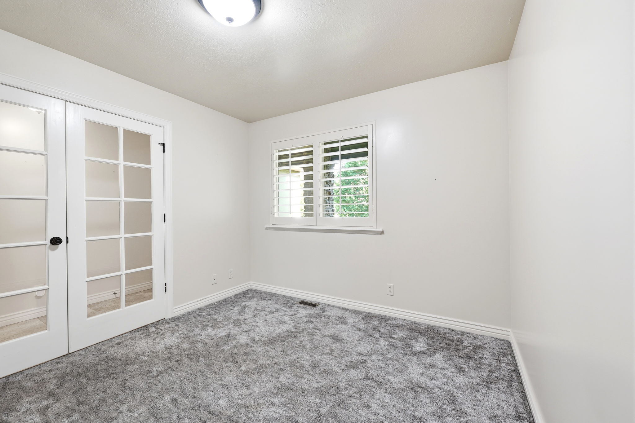 Room featuring double French doors with clear glass panes, a window with white plantation shutters, gray textured carpeting, white baseboards, and a flush-mount ceiling light fixture