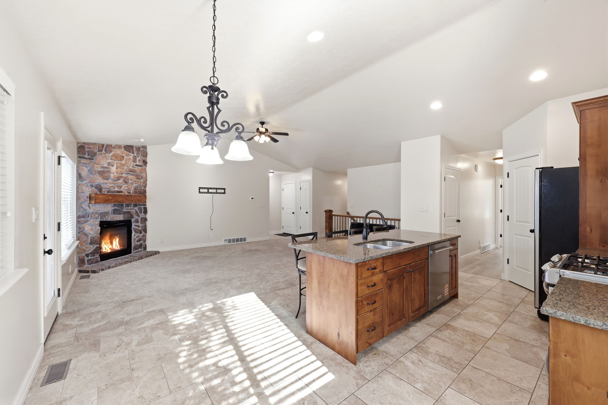 Spacious living area featuring a corner stone-faced fireplace with a wood mantel, an island kitchen with granite countertops and wood cabinetry, ceramic tile flooring, and recessed lighting
