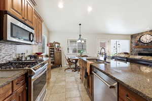 Kitchen featuring stainless steel appliances, dark stone counters, decorative light fixtures, backsplash, and a stone fireplace