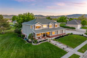 View of front of home at dusk featuring a large green lawn, flower beds, walkway to covered porch, surrounded by mountains
