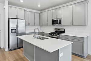 Kitchen with gray cabinets, stainless steel appliances, a kitchen island with sink, light wood-style flooring, and light stone counters