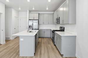 Kitchen with gray cabinetry, stainless steel appliances, a center island with sink, light wood-style floors, and light stone countertops
