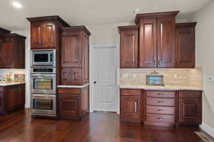Kitchen with backsplash, stainless steel appliances, light stone counters, dark wood-style flooring, and dark wood finish cabinets