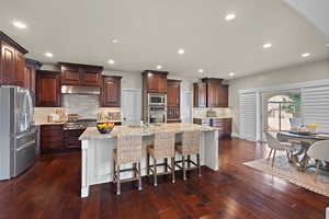 Kitchen featuring stainless steel appliances, a breakfast bar, light stone countertops, an island with sink, and dark wood finished floors