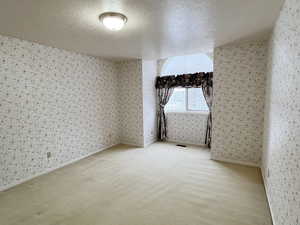 Empty room with a textured ceiling, light colored carpet, and wallpapered walls