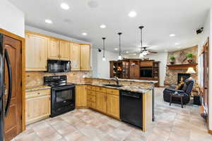 Kitchen featuring black appliances, open floor plan, light stone countertops, light wood finish cabinets, and a stone fireplace