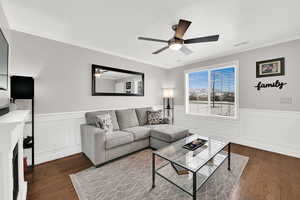 Living area featuring ornamental molding, a wainscoted wall, dark wood-style floors, ceiling fan, and a decorative wall