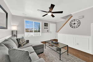 Living room featuring dark wood-style floors, crown molding, a ceiling fan, and a wainscoted wall