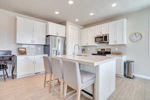 Kitchen with stainless steel appliances, white cabinets, light wood-type flooring, a kitchen breakfast bar, and a center island with sink
