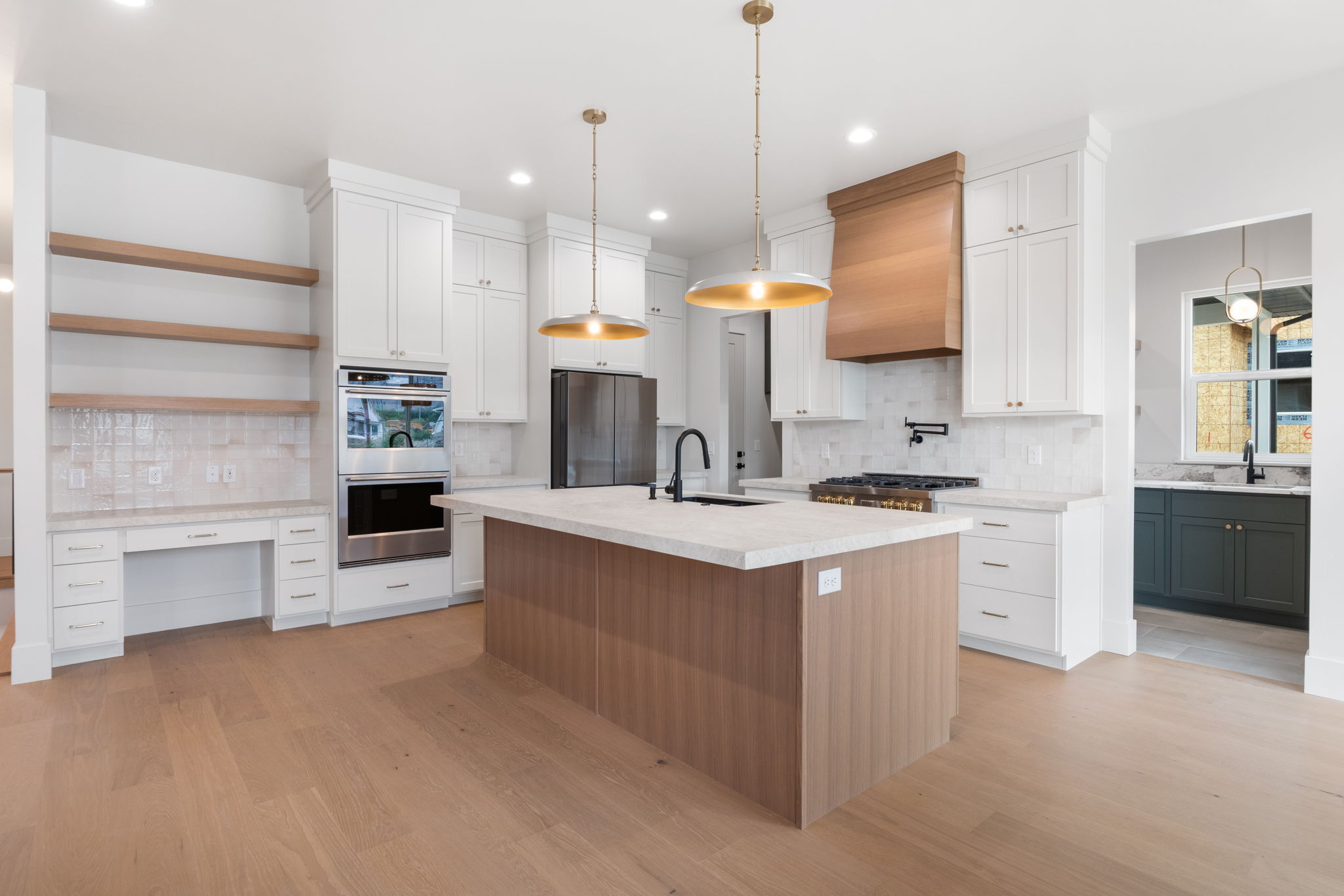 Kitchen featuring an expansive island with a light wood-finish base and light-toned countertop, white cabinetry with gold-toned hardware, stainless steel appliances, a wood-finished range hood, and wood-finish flooring