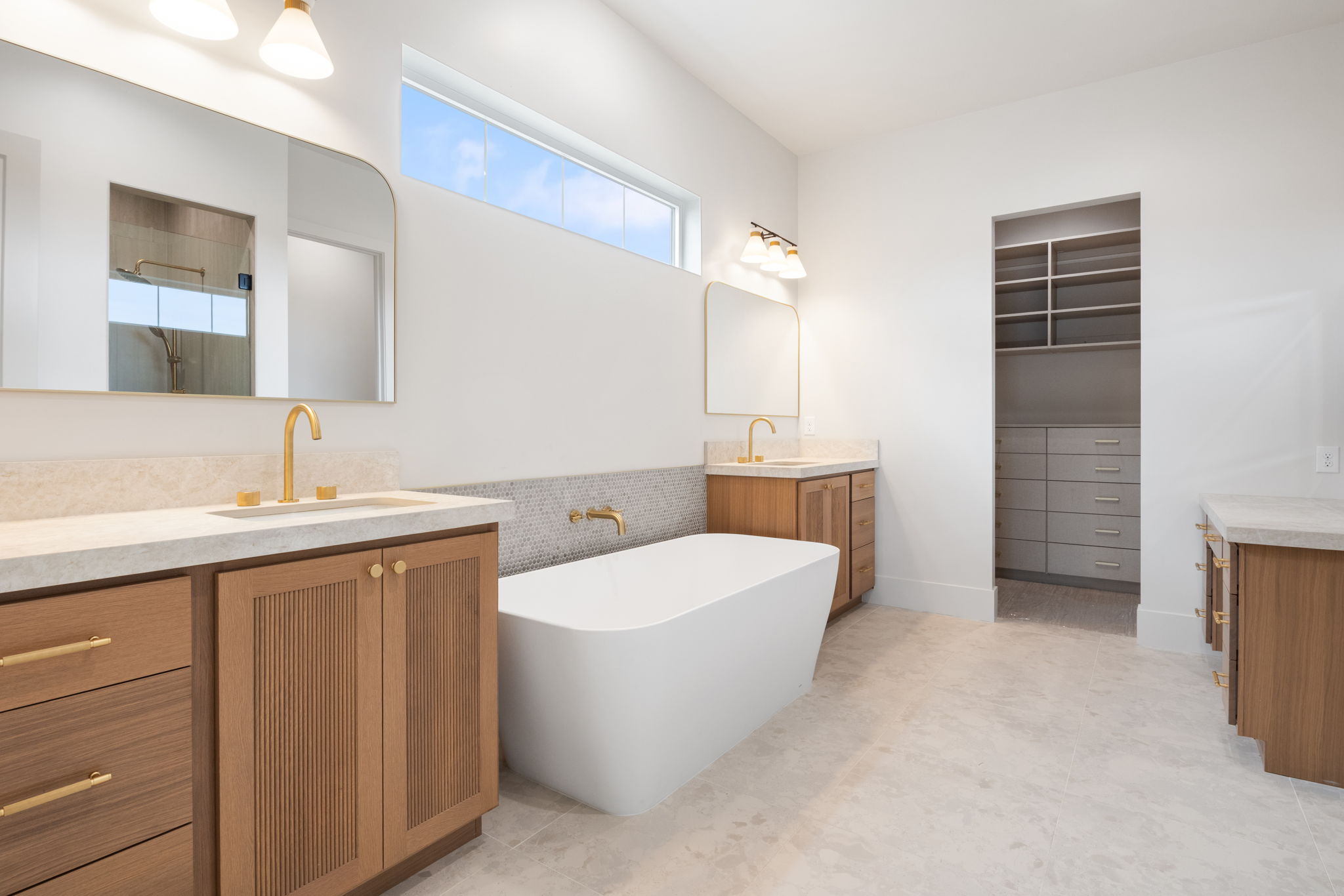 Contemporary bathroom featuring a freestanding soaking tub, dual wood-finish vanities with light countertops, and gold-tone fixtures