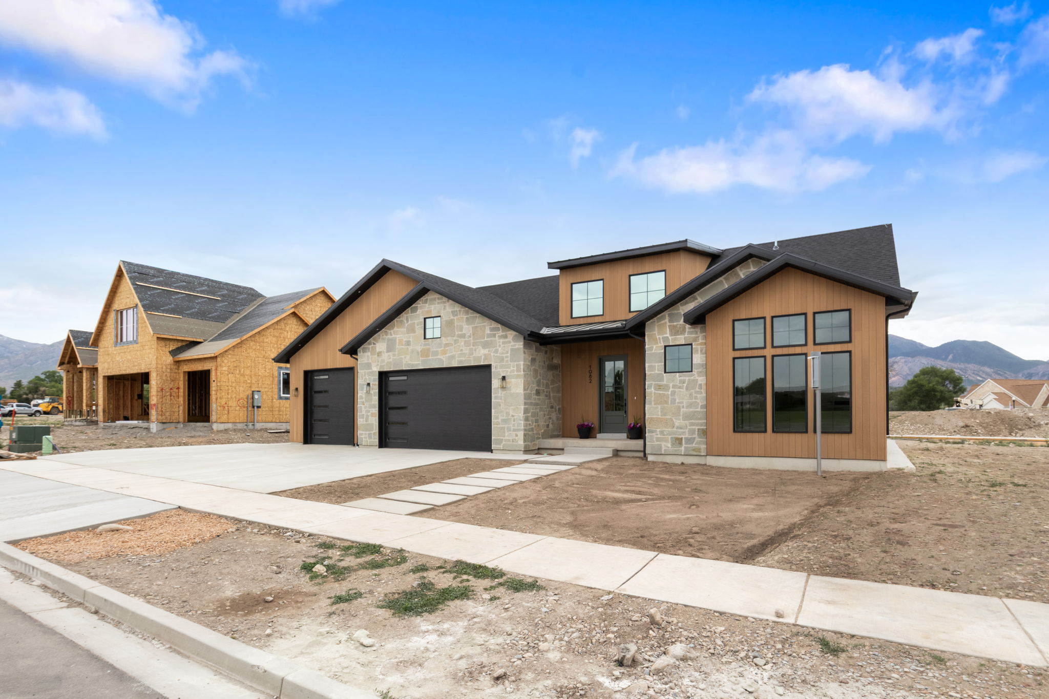 Modern exterior showcasing stone veneer and vertical wood-look siding, a black shingled roof, a two-car garage with contemporary black doors, and a concrete driveway