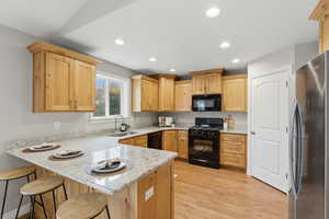 Kitchen featuring a peninsula, black appliances, a breakfast bar, light stone countertops, and light wood finish cabinets