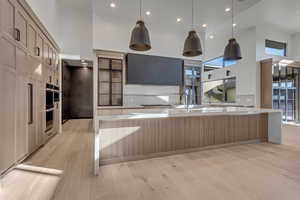 Kitchen with a high ceiling, a large island with sink, light wood-style flooring, light stone counters, and two tone cabinetry