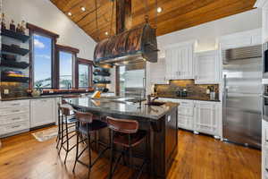 Kitchen with open shelves, backsplash, a vaulted wooden ceiling, an island with sink, and stainless steel appliances