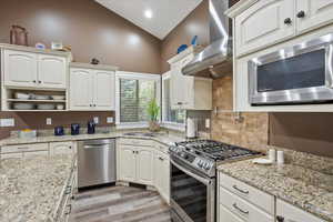 Kitchen with stainless steel appliances, open shelves, light stone counters, light wood-style flooring, and lofted ceiling