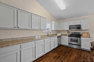 Kitchen featuring stainless steel appliances, light stone countertops, vaulted ceiling, white cabinets, and dark wood-style floors