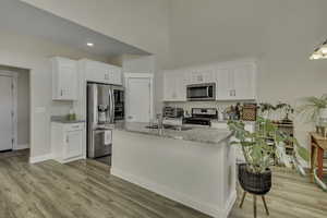 Kitchen with light stone counters, white cabinets, stainless steel appliances, an island with sink, and a high ceiling
