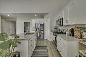 Kitchen featuring stainless steel appliances, white cabinetry, an island with sink, light stone counters, and light wood-type flooring