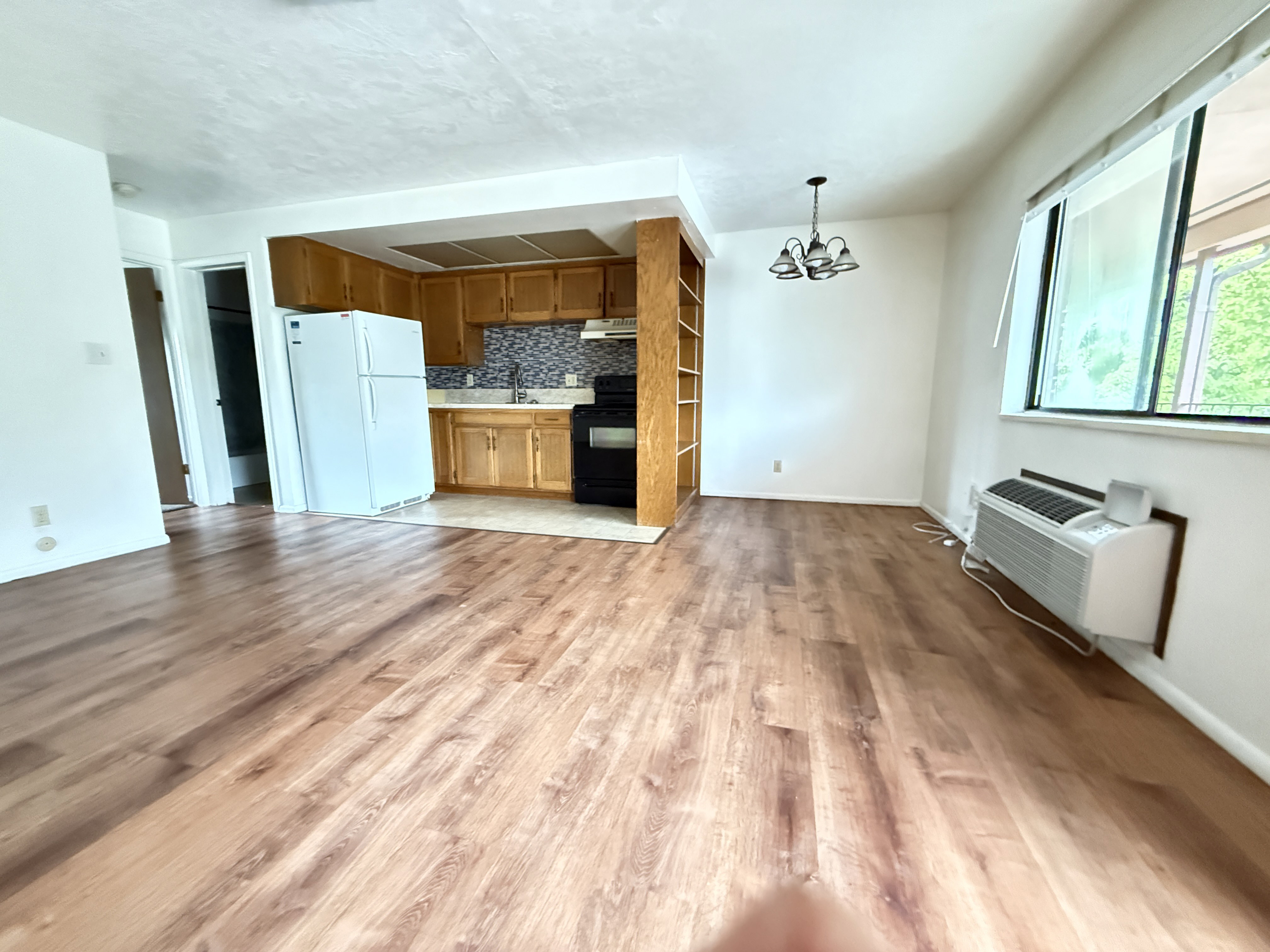 Open-concept living area featuring wood-finish flooring, a kitchen with light wood cabinetry and a tiled backsplash, and a built-in shelving unit