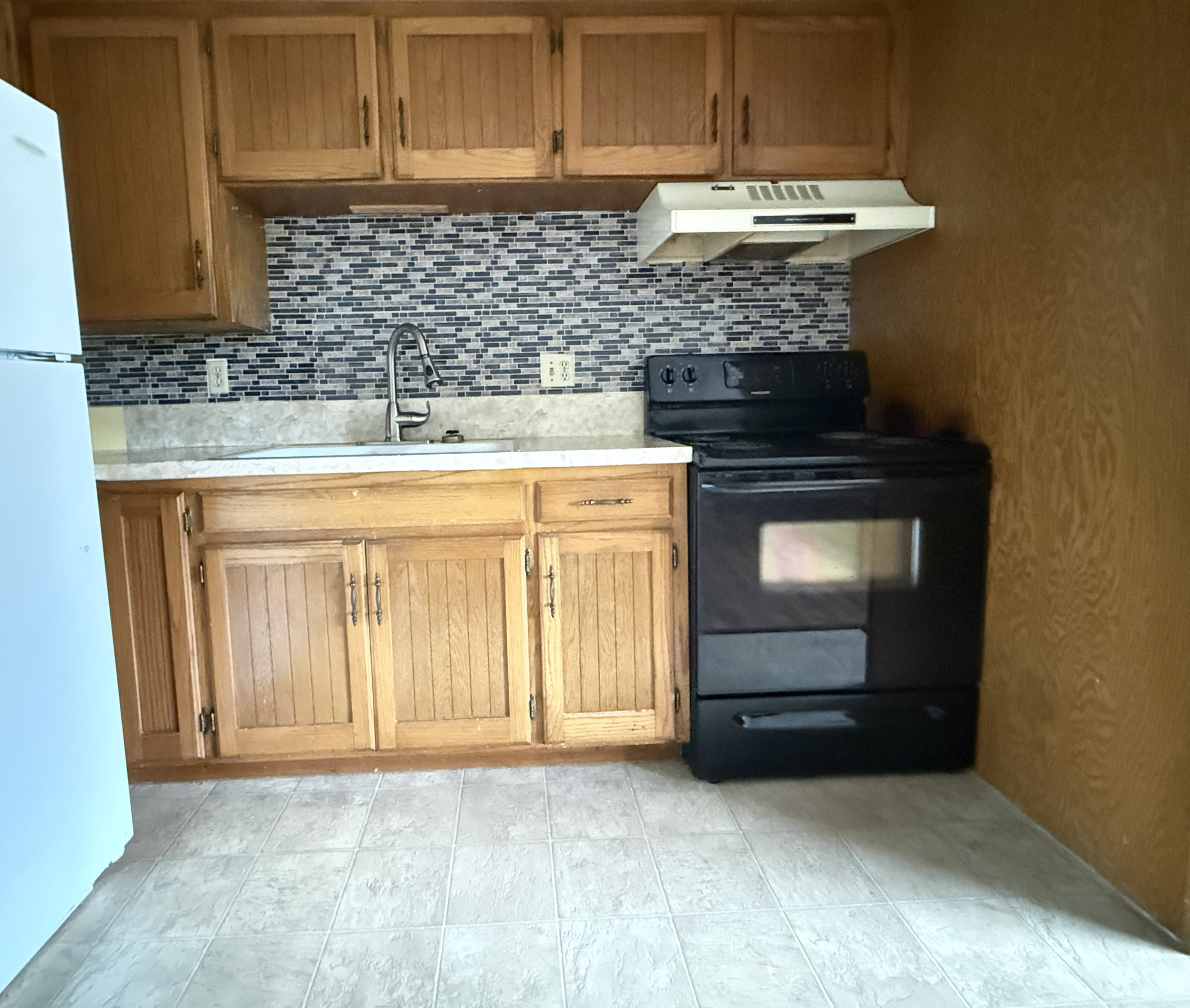 Kitchen featuring wood cabinetry, a mosaic tile backsplash, a stainless steel sink with a gooseneck faucet, light-toned countertops, and black appliances including a range and refrigerator