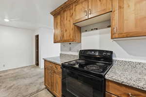 Kitchen featuring black electric range, light stone countertops, wood finish cabinets, and light colored carpet