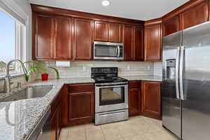 Kitchen featuring stainless steel appliances, decorative backsplash, light stone counters, light tile patterned floors, and bold wood finish cabinetry