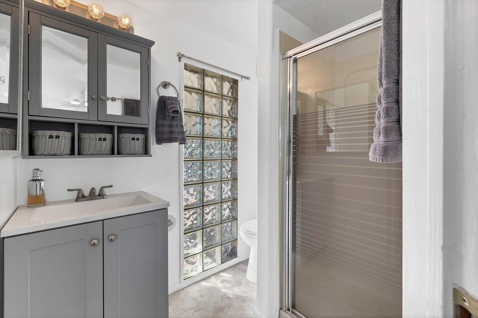 Bathroom featuring a gray vanity with an integrated sink, a matching medicine cabinet, glass block window, and a glass-enclosed shower
