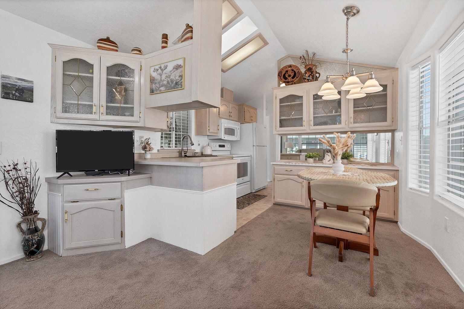 Open-concept kitchen and dining area featuring vaulted ceilings, skylights, white cabinetry with glass-front uppers, and an integrated dining nook with bay windows