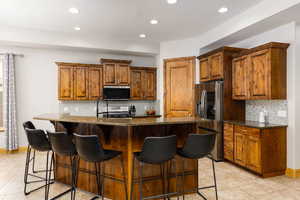 Kitchen featuring wood finish cabinetry, dark stone counters, stainless steel appliances, an island with sink, and recessed lighting