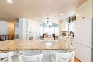 Kitchen with white appliances, a kitchen breakfast bar, light stone countertops, white cabinets, and a chandelier