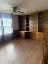 Unfurnished office featuring built in desk, wainscoting, a textured ceiling, a ceiling fan, and dark wood-type flooring