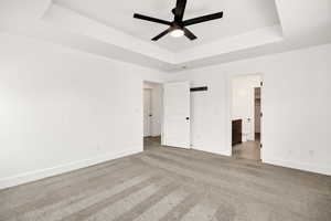 Unfurnished bedroom featuring a raised ceiling, ensuite bathroom, light carpet, and a ceiling fan