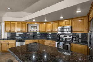 Kitchen featuring stainless steel appliances, wood finish cabinets, tasteful backsplash, dark stone counters, and open shelves