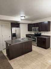 Kitchen featuring dark wood finish cabinets, a kitchen island with sink, stainless steel appliances, dark countertops, and a textured ceiling