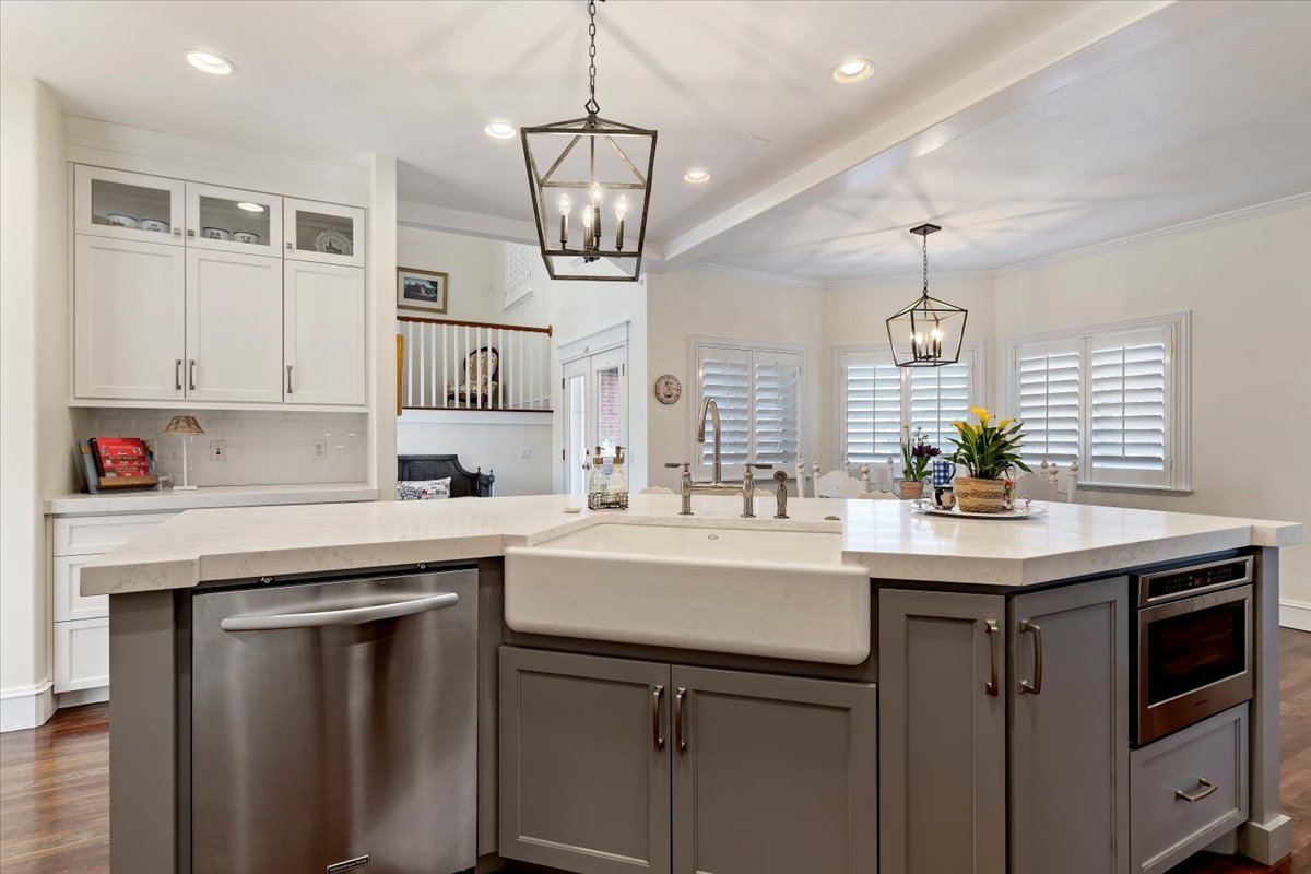 Dual tone kitchen with dark wood finished floors, suspended lighting, stainless steel dishwasher, glass fronted cabinets, and a center island with sink