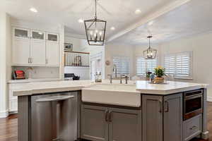 Dual tone kitchen with dark wood finished floors, suspended lighting, stainless steel dishwasher, glass fronted cabinets, and a center island with sink