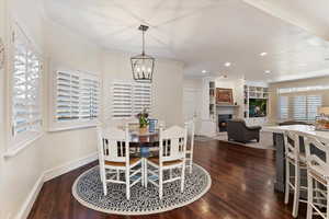 Dining area featuring a warm lit fireplace, dark wood-style flooring, suspended lighting, ornamental molding, and built in features