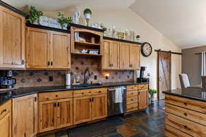 Kitchen with a barn door, dishwasher, lofted ceiling, dark stone countertops, and stone tile flooring
